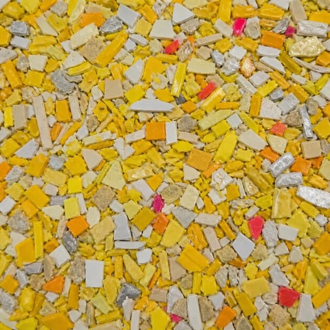Colorful animal feed or cereal mix with yellow corn, tan grain pieces, and scattered red and orange bits throughout